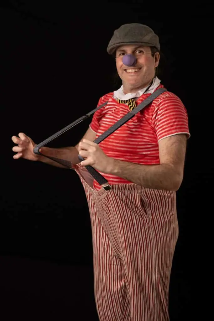 Francesco Scatarzi vestito da clown che sorride girato verso la telecamera, mentre si regge le bretelle dei pantaloni